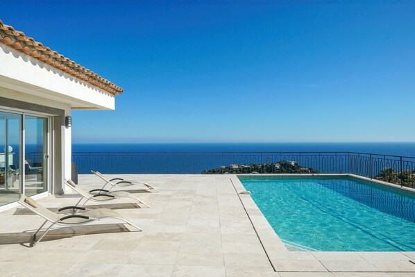 A pool area with a clear view of the ocean, lounge chairs, and a building with a tiled roof. - Modern villa in Californian style with breathtaking sea view (Roquebrune-sur-Argens)