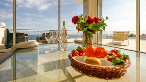 A glass table with a bottle of wine, two wine glasses, a vase with red roses, and a basket of fruit.