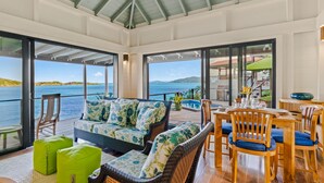A sunlit room with a large window overlooking a body of water, a wicker sofa, wooden chairs, and a dining table set for two. - Great House Villa, Jost Van Dyke (Jost Van Dyke)