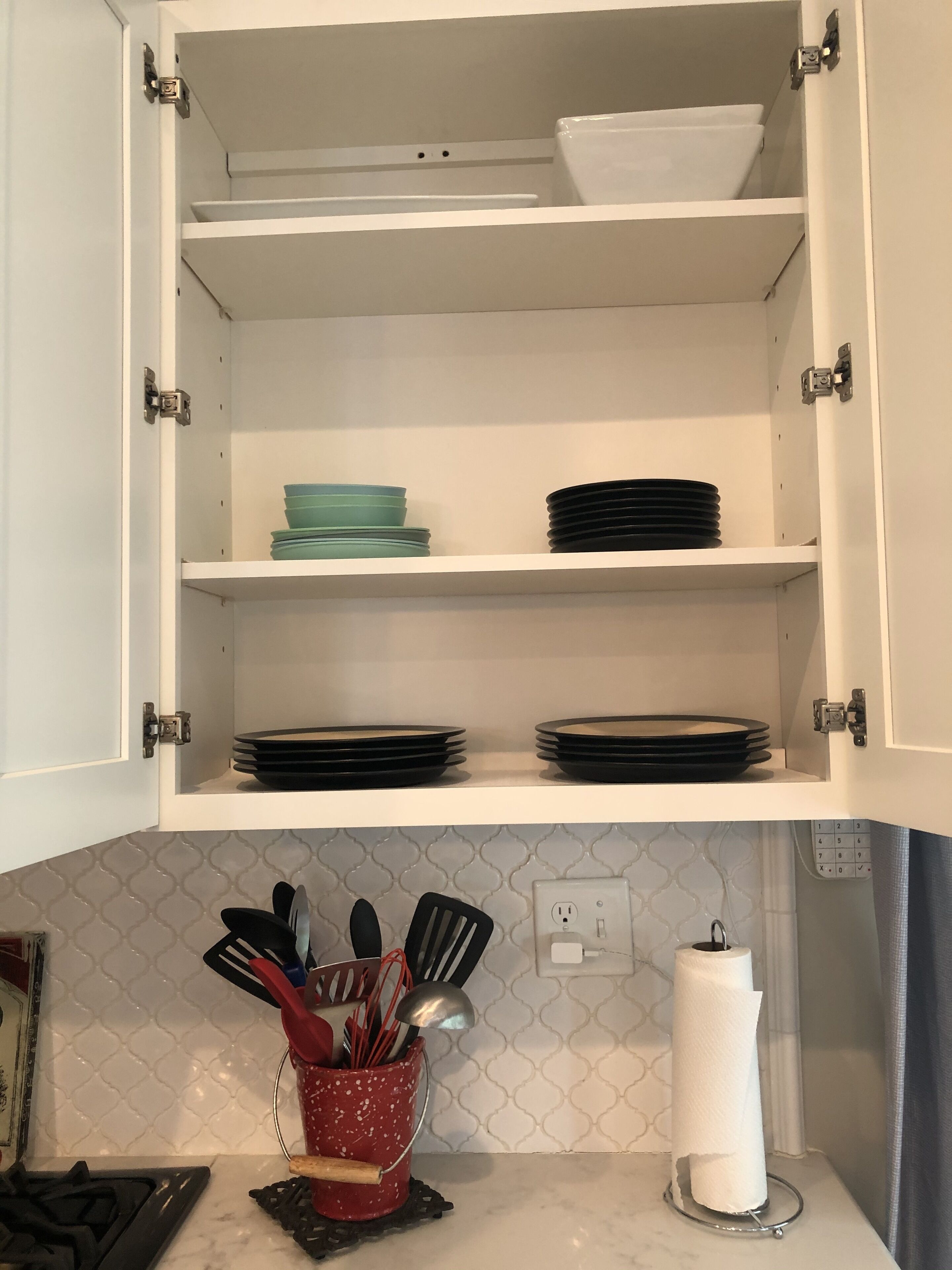 Un armario de cocina con estantes que contienen platos y tazones. Abajo, una encimera tiene un recipiente rojo para utensilios y un dispensador de toallas de papel.