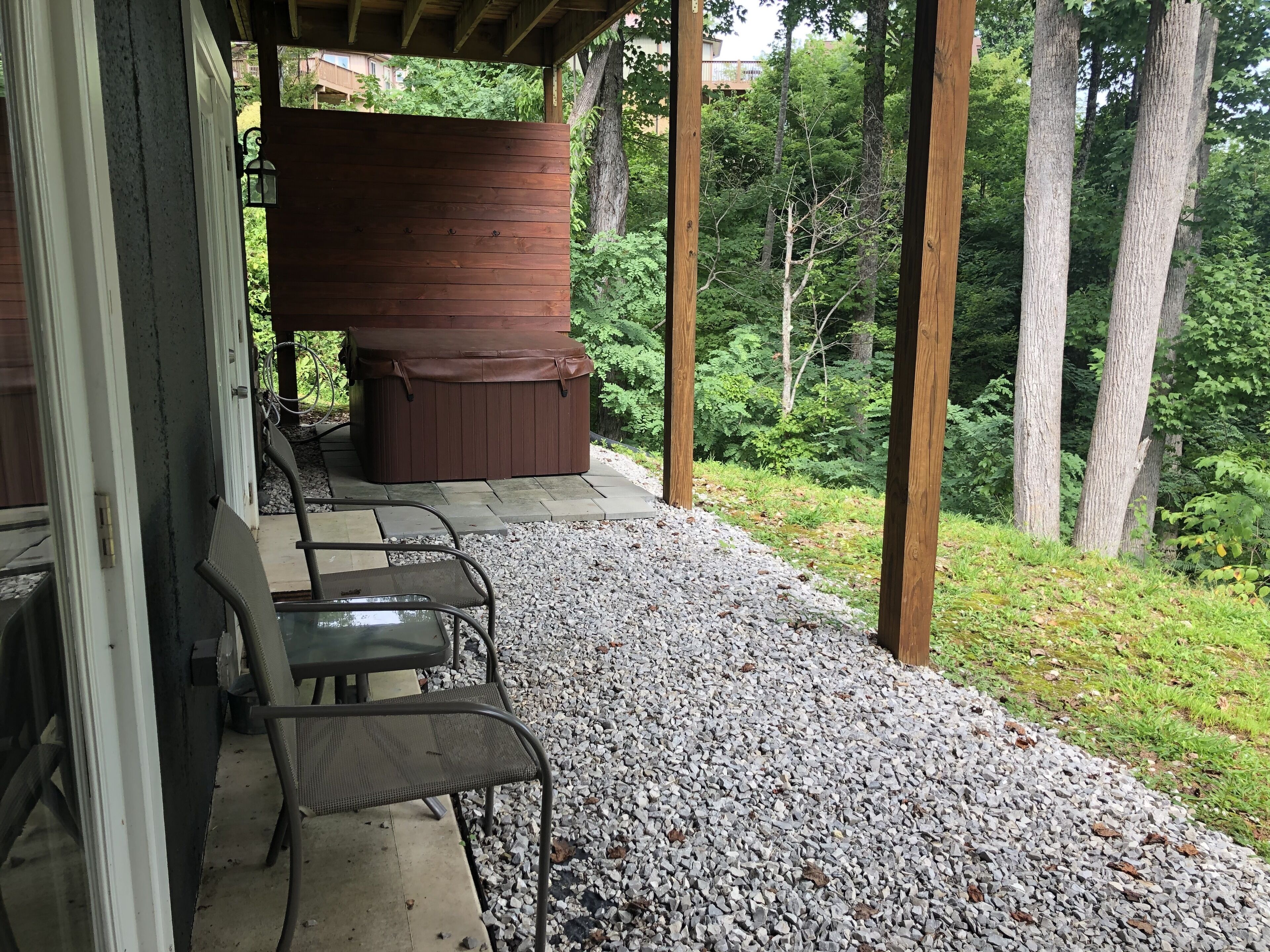 An outdoor seating area with a hot tub, surrounded by a wooden deck and gravel, overlooking a forested area.