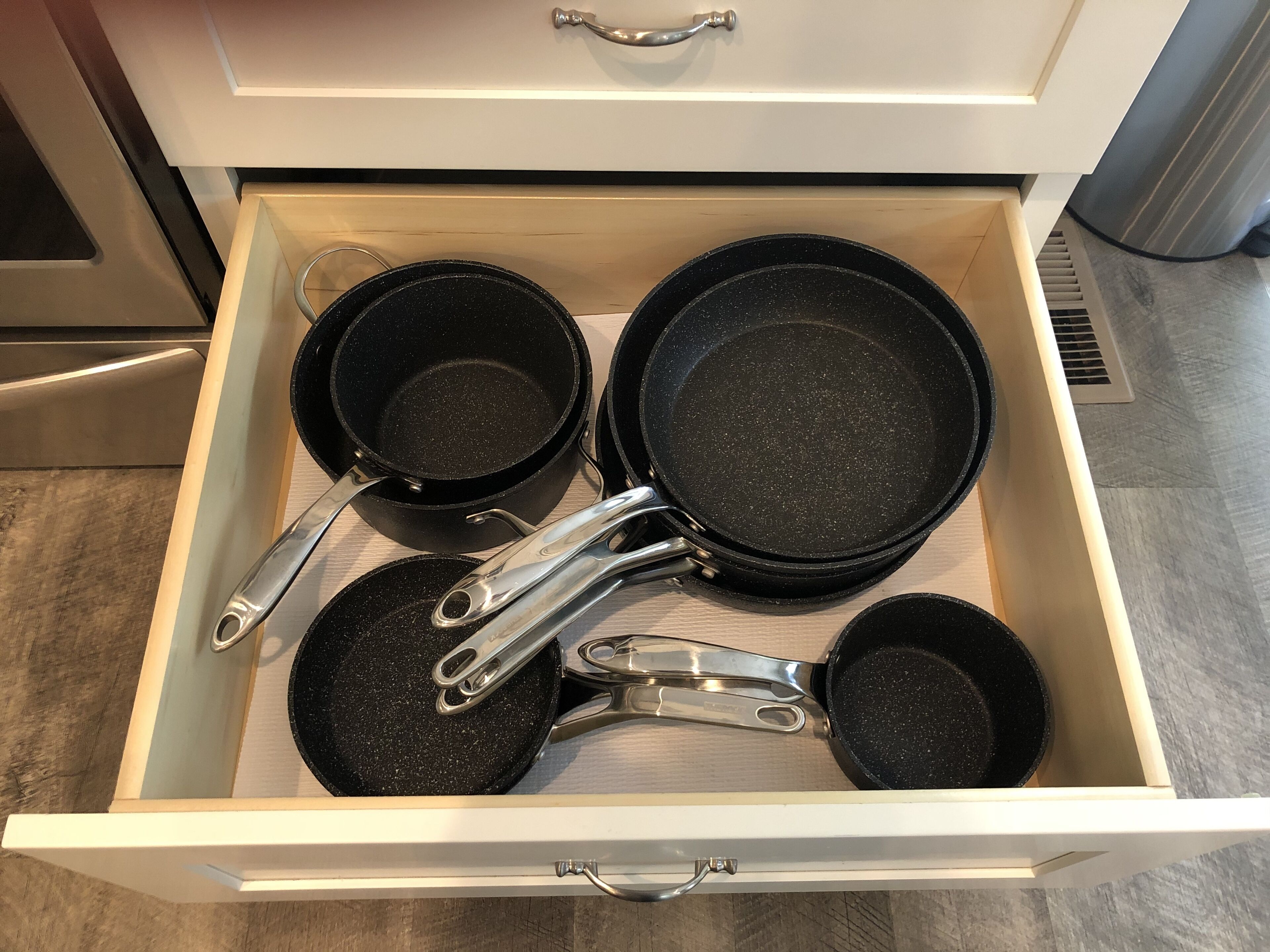 A drawer containing several black frying pans and a pair of metal tongs.