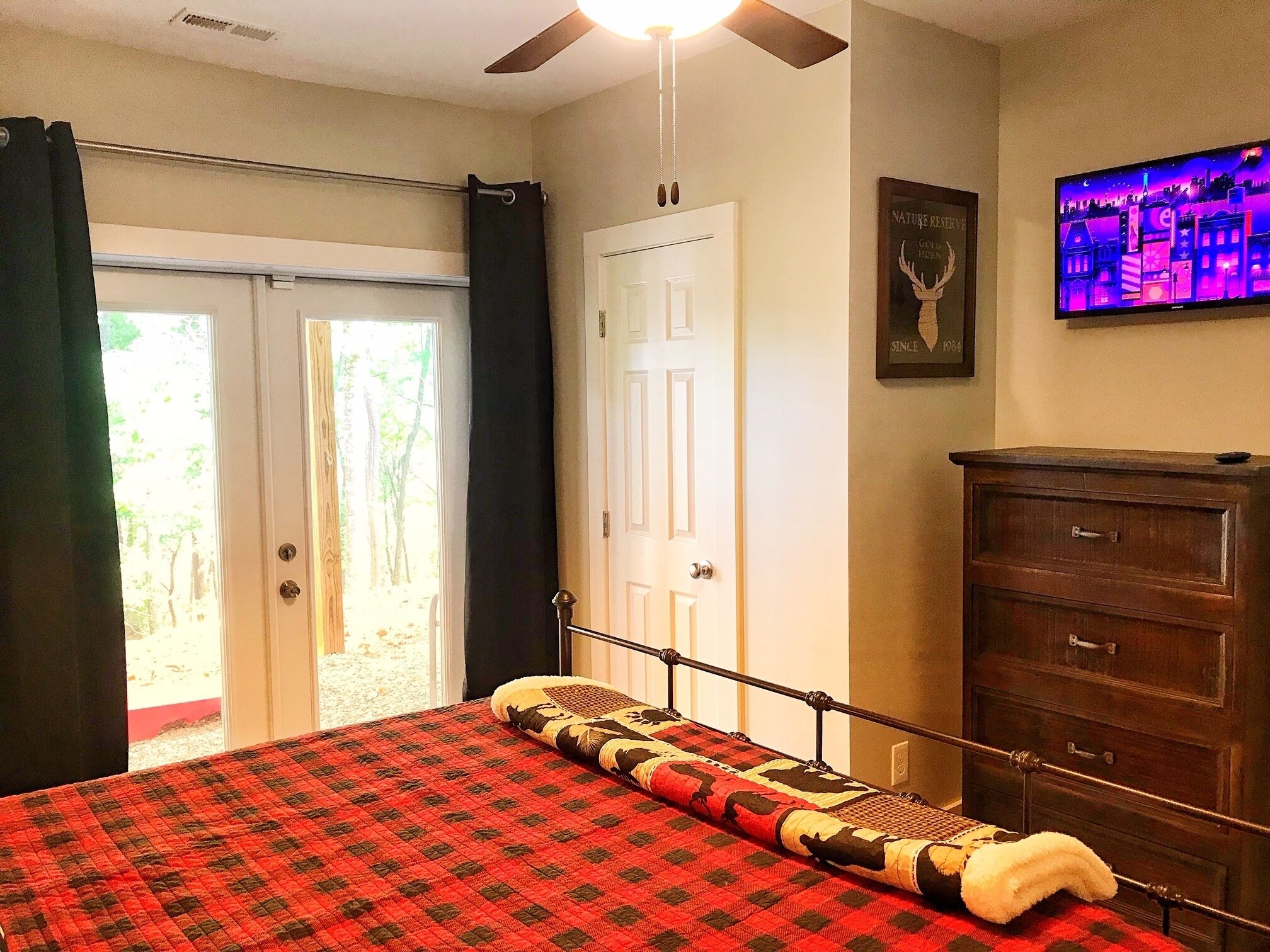 Un dormitorio con cama, cómoda, ventilador de techo, televisor y puerta que da a un balcón.