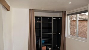A room with a built-in closet featuring multiple shelves and hanging space, a window with a view of a brick building, and a door with beige curtains. - Beautiful apartment located in the Louvre-Bollaert district (Lens)