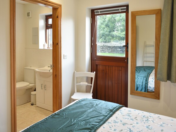 A bedroom with a bed, a wooden door, a white chair, a wooden wardrobe, and a bathroom visible through an open door.