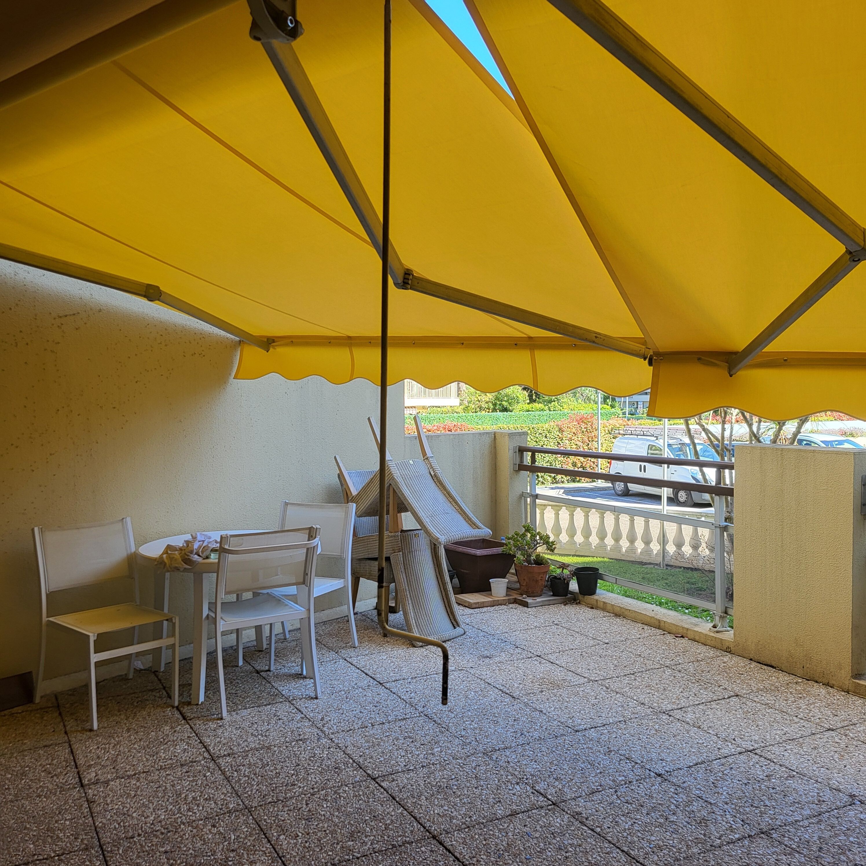 An outdoor patio with a yellow canopy, white chairs, a small table, and a potted plant.