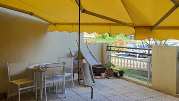 An outdoor patio with a yellow canopy, white chairs, a small table, and a potted plant.