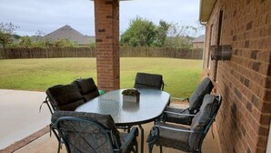 Een overdekt terras met een tafel met glazen blad en vier stoelen, een bakstenen muur en uitzicht op een gazon.