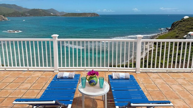 A balcony with two blue lounge chairs, a small table with a vase of flowers, and a view of the ocean and distant hills. - Caribbean Cottage with Breathtaking Views and Your Own Private Pool! (Cruz Bay)