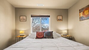 Un dormitorio con una cama grande, dos mesitas de noche con lámparas, un ventanal con vistas a árboles y decoración inspiradora en la pared.