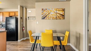 Un comedor con una mesa de vidrio y sillas amarillas, un refrigerador negro, armarios de madera y un extintor en la pared.