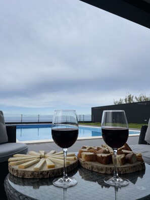 Two wine glasses on a table with cheese and bread, overlooking a swimming pool.