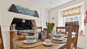 Een eetgedeelte met een houten tafel gedekt voor vier personen, omringd door stoelen. Achtergrond toont een keuken met een fornuis en oven.