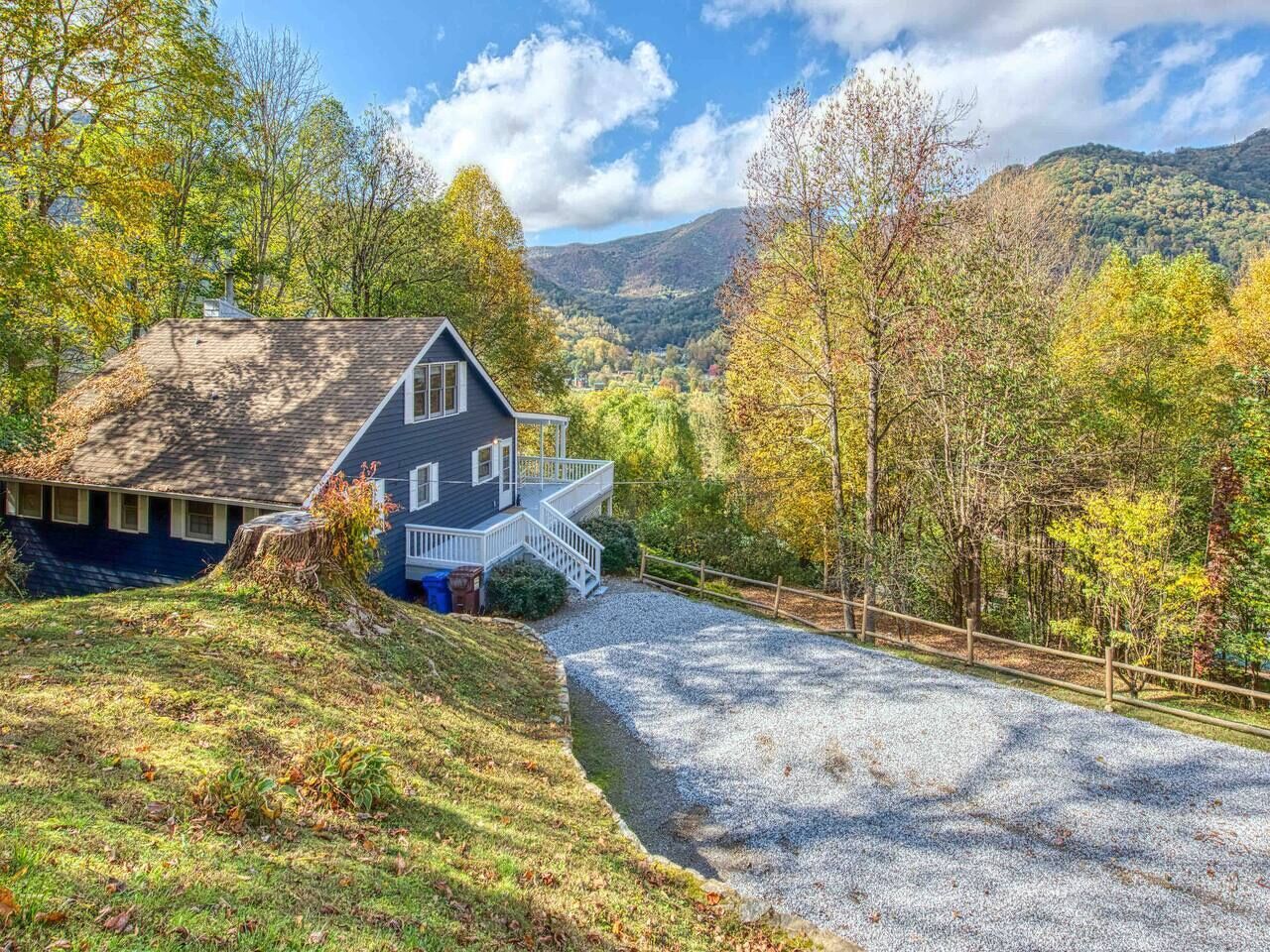 A house with a porch, surrounded by a gravel driveway and a wooden fence, set against a backdrop of autumn foliage and hills.