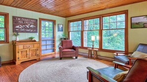 Ein gemütliches Zimmer mit Holzbalkendecke und großen Fenstern, einem Holzschrank, einem roten Sessel und einem runden Teppich.