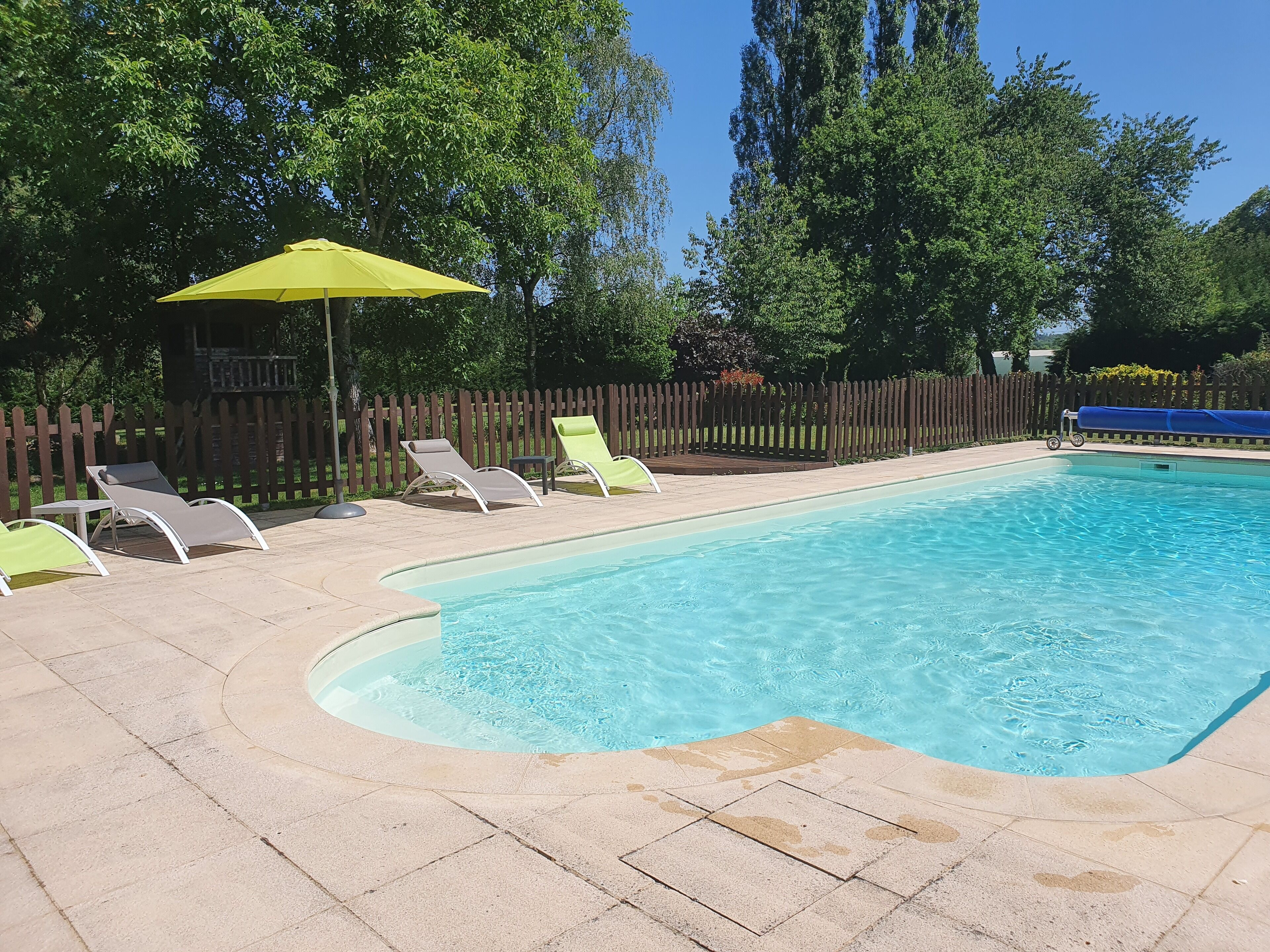 Une piscine rectangulaire à l’eau d’un bleu limpide, entourée d’une terrasse carrelée. On y trouve trois chaises longues et un parasol jaune près de la piscine.