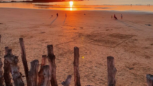 Una playa con un atardecer, postes de madera en primer plano y una estructura distante en el horizonte.