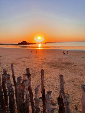 A beach with a sunset, wooden posts in the foreground, and a distant structure on the horizon.