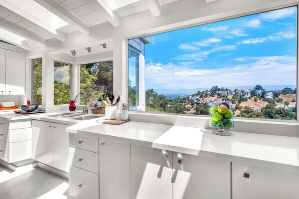 A modern kitchen with large windows, a view of a hillside, and a fruit bowl on the counter.