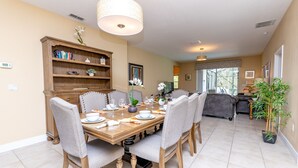 Un coin repas avec une table en bois dressée pour six personnes, entourée de chaises à revêtement gris. Une bibliothèque en bois, contenant divers objets, se trouve à gauche.