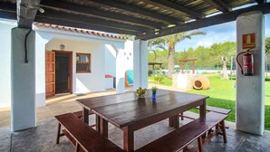 Un portico coperto con un tavolo e panche di legno, un estintore e vista su un giardino con una palma e un'area giochi.