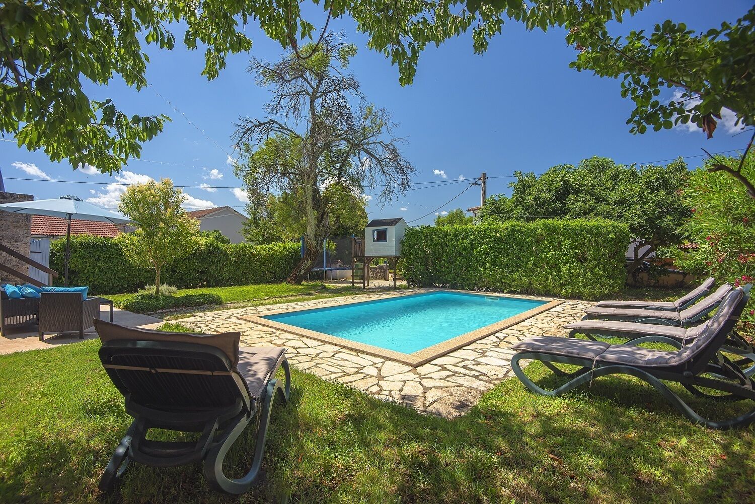 A rectangular swimming pool with a stone border, surrounded by grass and trees. There are two lounge chairs and a single chair facing the pool.