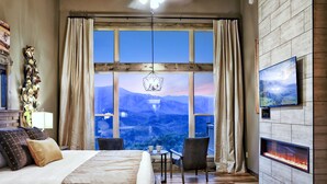 A bedroom with a large window, a bed, a chair, a small table, and a fireplace.