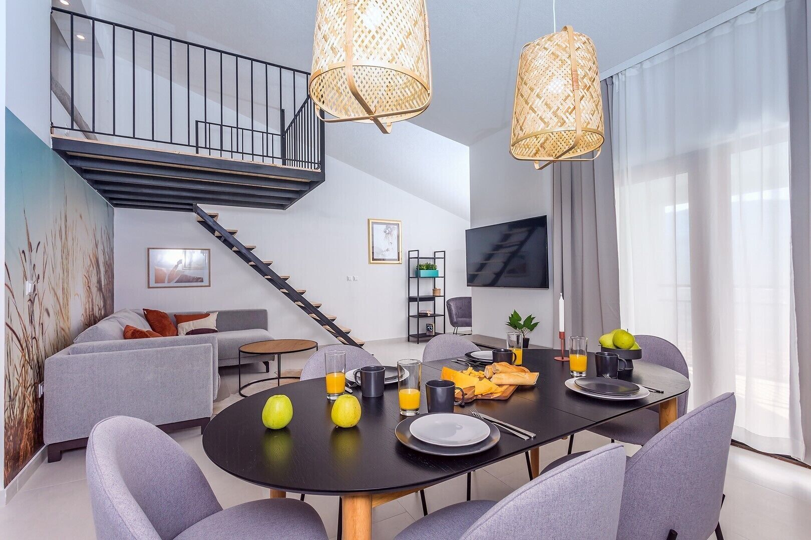 A modern living room with a dining area, featuring a staircase, a flat-screen TV, and a large window with sheer curtains.