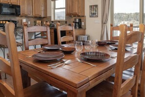 A dining table set with plates, bowls, and cutlery, surrounded by wooden chairs in a kitchen with wooden cabinets and a view of a snowy landscape.