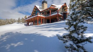 A large, multi-story log cabin with a prominent chimney, surrounded by snow-covered trees and a clear sky.