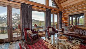 A log cabin interior with a large glass table, leather sofas, and a view of snow-capped mountains.
