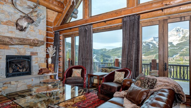 A log cabin interior with a stone fireplace, leather sofa, and a view of mountains through large windows.