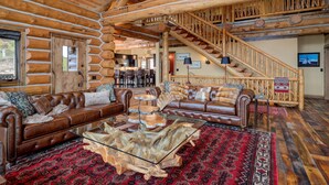 A log cabin interior with leather sofas, a glass coffee table, and a wooden staircase.