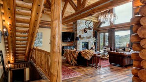 A log cabin interior with a stone fireplace, wooden beams, a fireplace mantel with antlers, and a view of the outdoors through large windows.