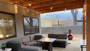 An outdoor patio with wicker furniture, a table with a fire feature, and a view of a brick wall and buildings in the background. - Private house in a safe area, close to Nob Hill, UNM, Airport, Netflix, and KAFB (Albuquerque)