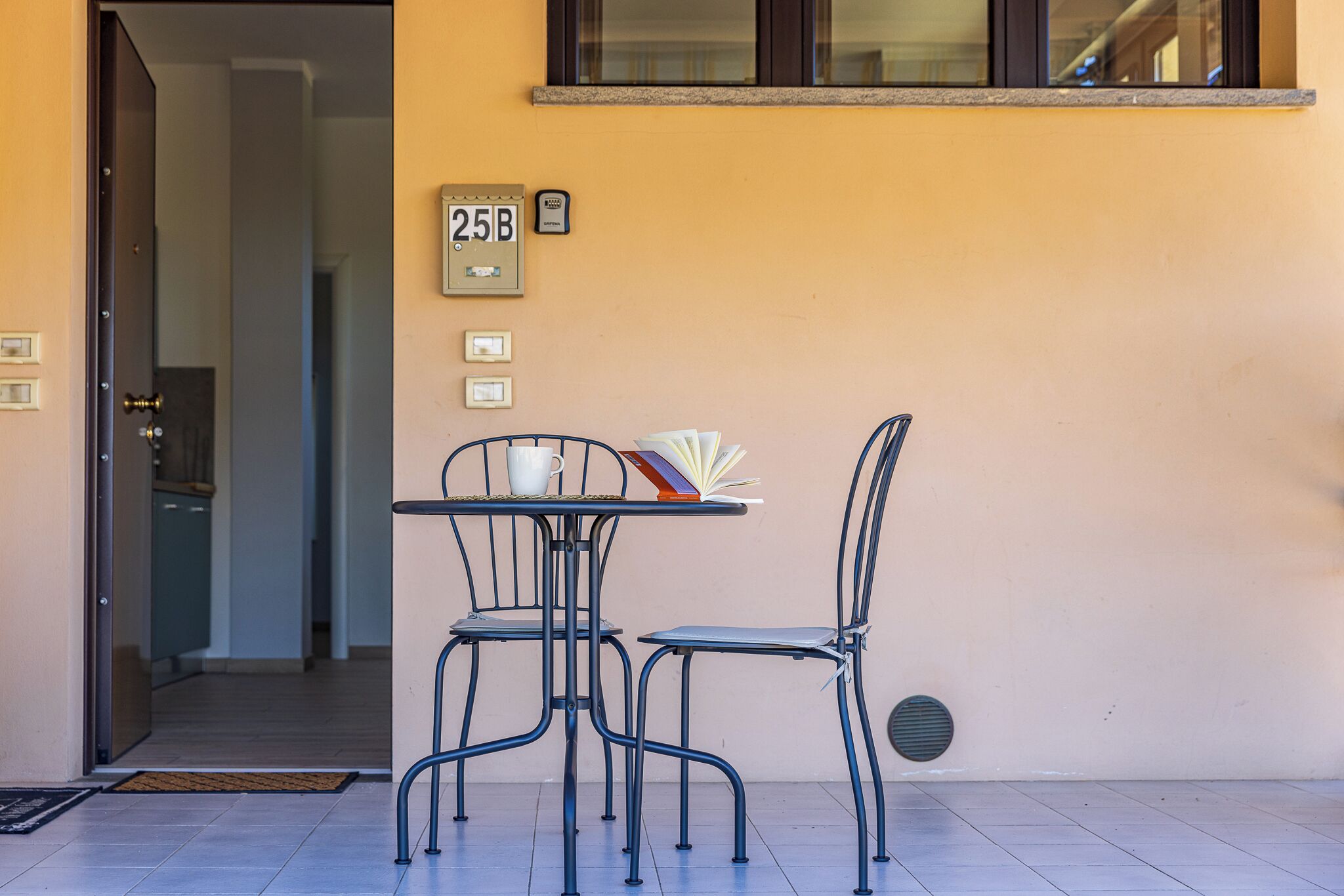 Un espace extérieur aménagé avec une table et deux chaises, une porte donnant sur l’intérieur et une boîte aux lettres portant le numéro 25B sur le mur.