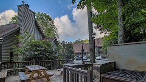 Una terraza de madera con mesas de picnic y zona de barbacoa, rodeada de árboles y un edificio con chimenea.
