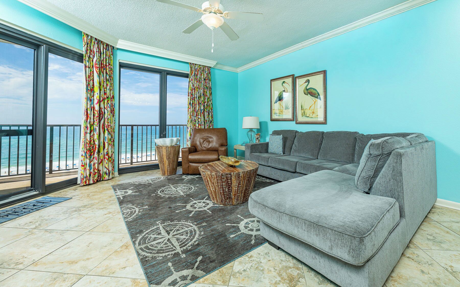 A living room with a grey sofa, a brown armchair, a round coffee table, and a large window overlooking the beach.