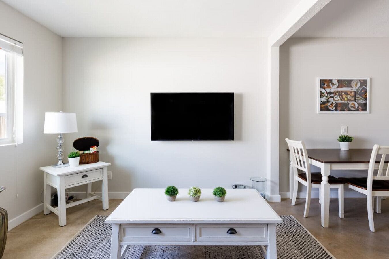 A modern living room with a white sofa, a coffee table, a TV mounted on the wall, and a dining area with a table and chairs.