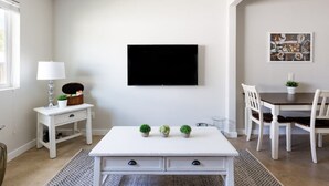 A modern living room with a white sofa, a coffee table, a TV mounted on the wall, and a dining area with a table and chairs. - Stylish 2 Bedroom Camelback (Phoenix)