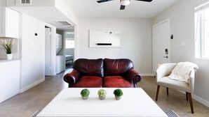 A modern living room with a dark red leather sofa, a white coffee table with three small potted plants, a ceiling fan, and a white armchair with a blanket. - Stylish 2 Bedroom Camelback (Phoenix)