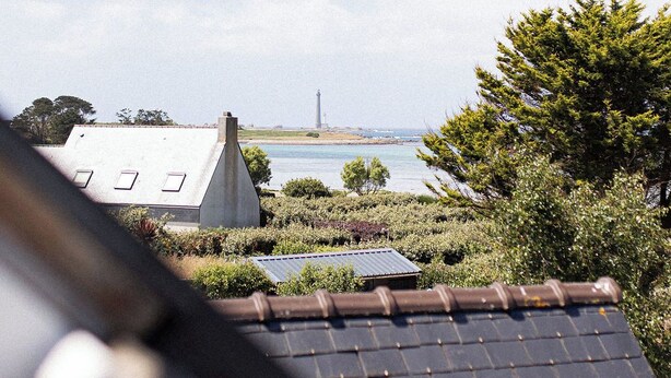 A view of a coastal area with a lighthouse in the distance, a house with a sloped roof, and a body of water. - Ty Glaz Lilia (Plouguerneau)