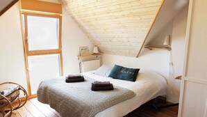 A bedroom with a sloped ceiling, a bed with a white comforter and a dark pillow, a wooden floor, a window, and a radiator. - Ty Glaz Lilia (Plouguerneau)