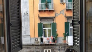 Vista attraverso una finestra su un edificio con facciata gialla, persiane verdi e un piccolo balcone con ringhiera in metallo.