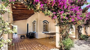 A covered outdoor patio with a table and chairs, a wooden pergola, and a wall with arched windows.