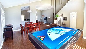 Een ruime woonkamer met een pooltafel, eettafel en stoelen. Een trap leidt naar een hoger gelegen verdieping.