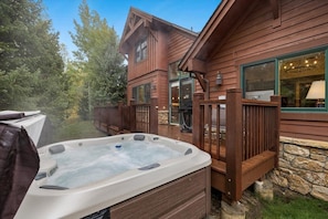 Un jacuzzi al aire libre frente a una cabaña de madera con base de piedra, rodeado por una terraza de madera y un muro de piedra.