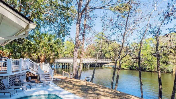 Un espace piscine doté d’une terrasse, de chaises longues et offrant une vue sur une rivière bordée d’arbres et traversée par un pont.