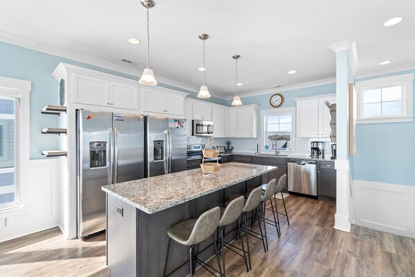 A modern kitchen with white cabinets, stainless steel appliances, a granite countertop, and a large island with bar stools. - Just Add Water - Spacious 7BR oceanfront home in Oak Island, perfect for 19 guests. (Oak Island)