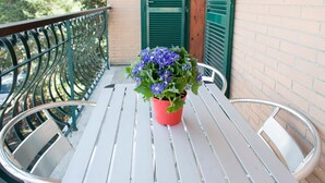 Ein Balkon mit Metallgeländer, einem Blumentopf mit blauen Blüten und einem weißen Tisch mit Metallstühlen.